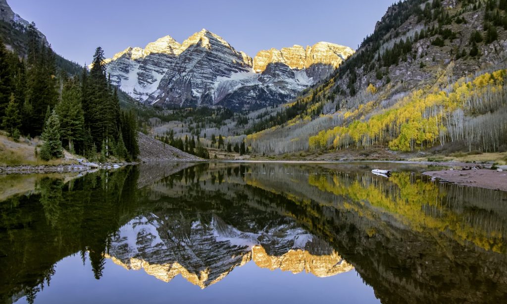 Sunrise over the iconic Maroon Bells mountains near Aspen, Colorado.  Clear sky above with colorful aspen trees in full fall foliage color splendor with the entire scene reflected in the small lake in the foreground.