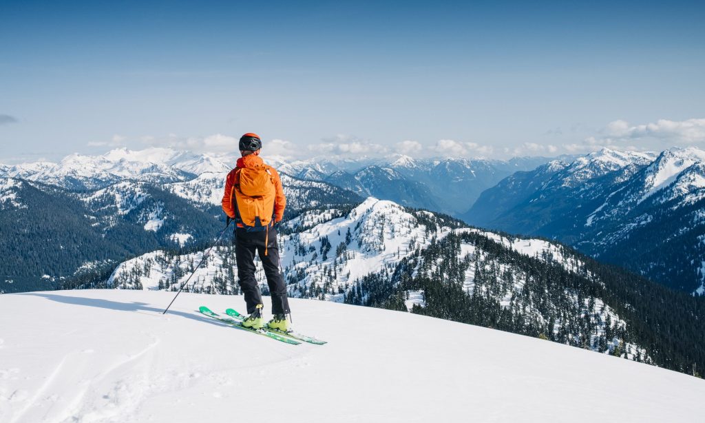 April, 05, 2021 - Squamish, BC, CAN: A male ski mountaineer stands on top of Anif mountain and looks out at a dramatic view of the surrounding mountains and deep valleys far bellow.