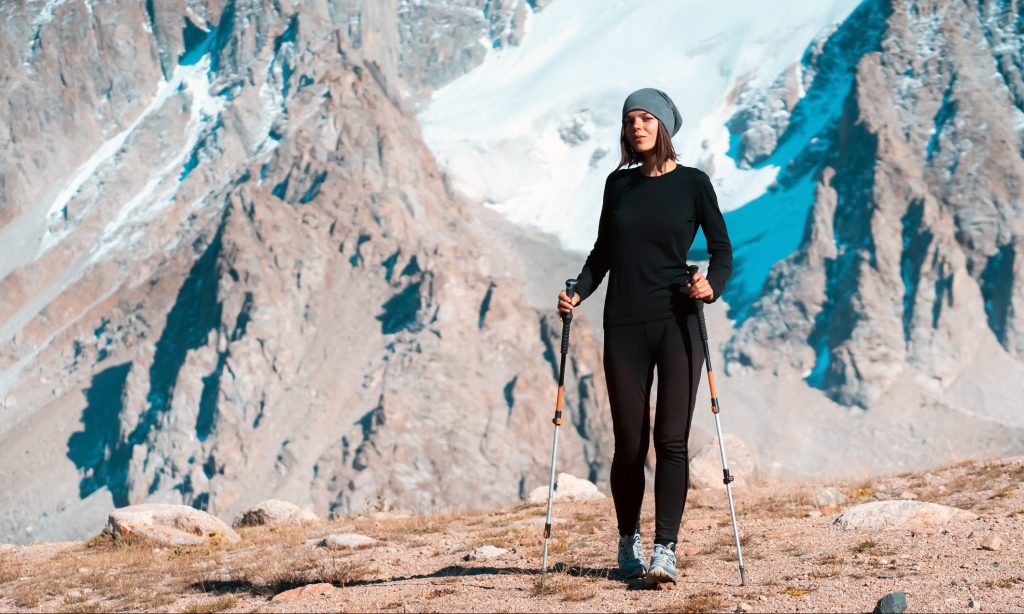 A young beautiful sports girl in thermal underwear with trekking poles walks along the trail among the snow-capped high peaks of the mountains, the traveler walks and climbs tin the national park.