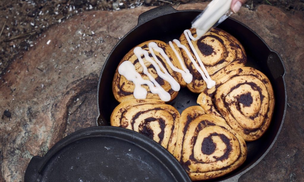 Hand pouring icing over cast iron made cinnamon rolls on a boulder.