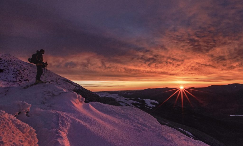 skier stands on mountain in winter with stunning red pink sunrise