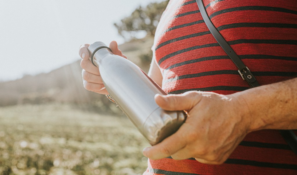 A man stands outside in a sunny environment and holds a silver coloured metallic reusable flask / water bottle. Space for copy.