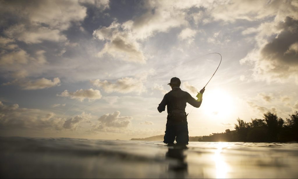 A man fly fishing under a dramatic sunrise over the Pacific Ocean.